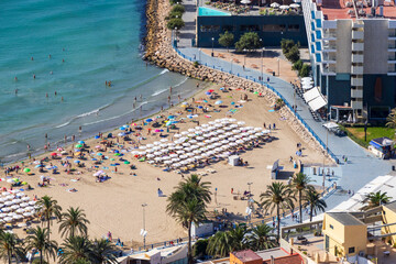 Aerial view of a vibrant beach in Alicante, featuring colorful umbrellas and sunbathers enjoying the sunny day. The blue sea complements the sandy shore, creating a perfect summer getaway.