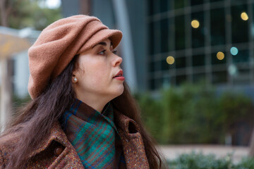 Portrait of a young Caucasian woman in a hat and scarf standing outdoors near a glass building....