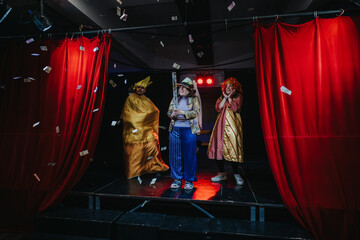 A lively theatrical performance featuring three actors dressed in vibrant costumes, surrounded by...