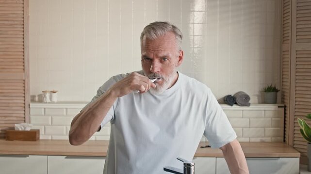 Aged man with grey beard brushing teeth with electric toothbrush in front of mirror in bathroom and spitting toothpaste into sink during daily hygiene ritual