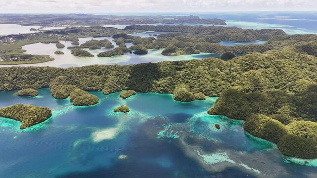 Scenic drone shot sweeping across the vivid aqua ocean and green-covered terrain with a small town in the distance in Palau. Wide shot of town and islands