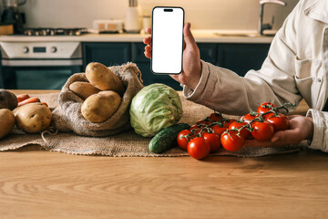 Person holding a smartphone with blank white screen over a rustic kitchen table filled with fresh...