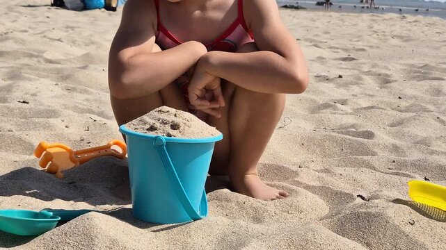 A young child plays on a sunny beach, building with a blue bucket filled with sand. Captured during summer vacation, this moment reflects carefree joy and early childhood exploration.