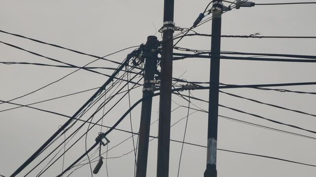 A cluttered view of tangled and messy electrical and telephone cables strung across poles in a dense urban environment. This disorganized wiring setup is common in many developing cities and highlight