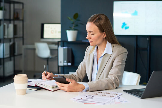 Manager at office desk using smartphone and writing notes