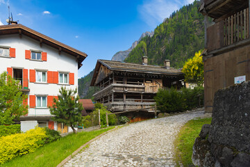 A charming cobblestone street in Sottoguda, a traditional Alpine village, flanked by historic wooden and stucco houses with lush green mountains in the background.