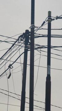 Portrait-format video showing a chaotic tangle of electrical and telephone cables hanging across utility poles in an urban neighborhood. The disorganized wiring system represents infrastructure