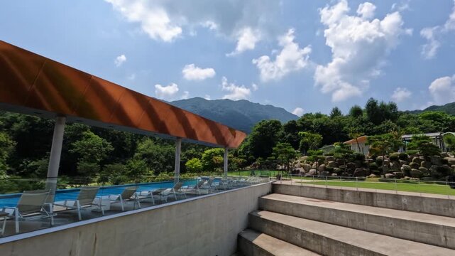 A timelapse captures beautiful moving clouds above a luxury resort swimming pool with a modern canopy and architectural concrete steps, set against a green mountain in Gapyeong.