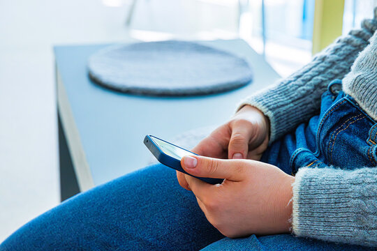 Closeup of a young person in a gray knit sweater and blue jeans using smartphone while sitting on a bench in a bright indoor space. 
