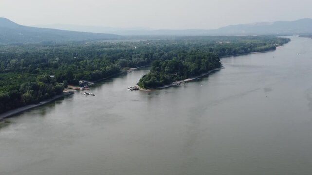 Drone video, Danube River near Budapest winding through a lush, forested region with a sandy beach and distant hills. Boats on the water, small riverside community stretches along the shoreline. 