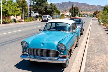 Ein leuchtend türkises Oldtimer-Auto mit weißem Dach parkt am Straßenrand einer sonnigen Straße. Im Hintergrund sind weitere Autos, Häuser, Bäume und Berge zu sehen