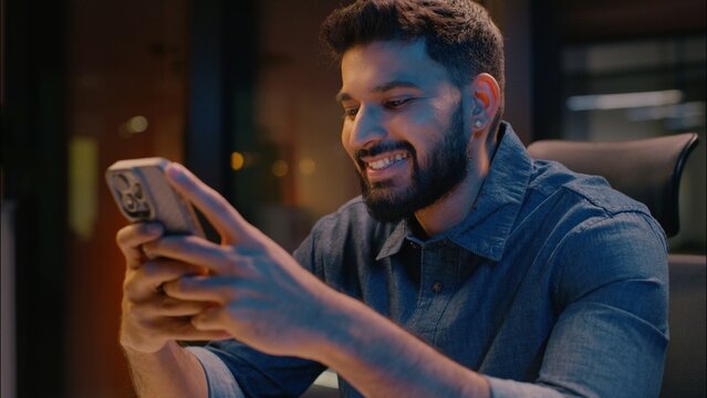 Indian ethnic businessman man business guy in evening office late night addict using mobile phone messaging scrolling gadget smartphone online chatting male employer manager browsing cellphone smiling