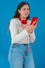 Concerned young woman in white sweater holding red smartphone with headphones, reacting in shock...