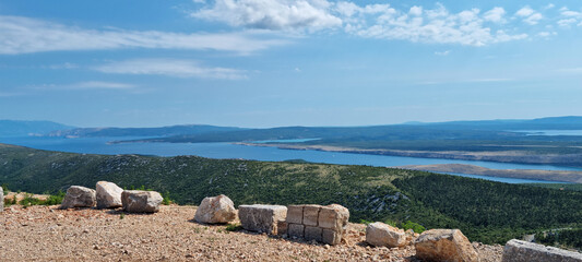 An expansive view from a viewpoint above Krizisce towards Krk Island, featuring land, sea, and islands under a blue sky with clouds
