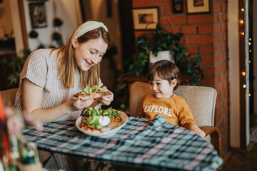 A cheerful mother interacts with her young child during a meal. Captured indoors in a warm home setting. Scene showcases love, motherhood, and shared moments of joy.