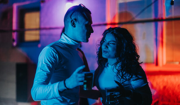 A moment shared between two friends laughing and conversing while holding drinks, set against a vibrant, neon-lit nightlife environment. - Powered by Adobe