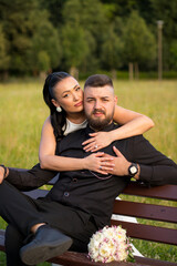 Bride hugging groom from behind, groom sitting on park bench. Atmospheric photo with emphasis on...