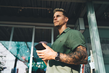 Millennial man with tech watch and smoothie holds smartphone in sunlight, capturing digital...