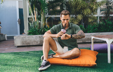 Tattooed man sipping smoothie while checking smartwatch, symbolizing digital mindfulness,...