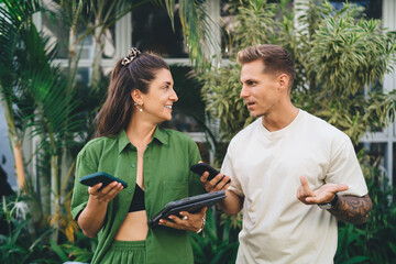 Freelance team having tech-focused conversation outside, both holding phones and tablet, reflecting...