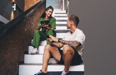 Man and woman seated on modern staircase, holding smartphone and tablet, engaged in relaxed...