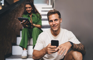 Confident man in foreground smiling with smartphone, woman behind working on tablet, showing dual...