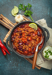 Traditional mexican chili con carne served in a cast iron pot, garnished with lime and red chili peppers, alongside sour cream and fresh ingredients on a rustic wooden board