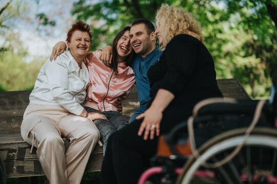Group of diverse individuals sitting together on a bench, laughing and embracing in a park setting, highlighting inclusion, friendship, and happiness under the beauty of nature. - Powered by Adobe
