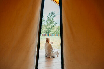woman in blanket sitting on wooden terrace and holding mug viewed from inside tent, peaceful...