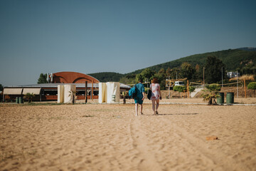 Two individuals stroll along a sandy beach under clear skies, enjoying a calm and scenic setting. A...