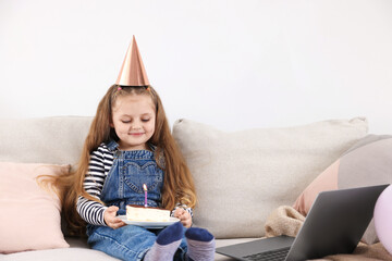 Birthday celebration. Little girl with piece of cake having video call via laptop on sofa at home