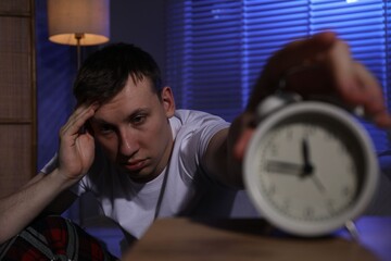 Frustrated man suffering from insomnia and turning off alarm clock indoors, selective focus