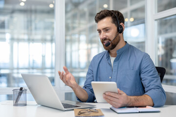 Fototapeta premium A man in a headset gestures while holding a tablet, sitting at a desk with a laptop, glasses, and notebooks.