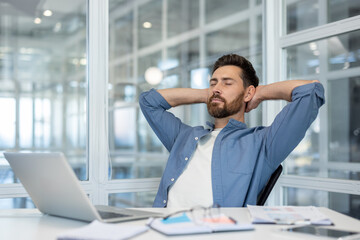 A relaxed businessman sits at his desk, taking a break with his hands behind his head. He is in his...