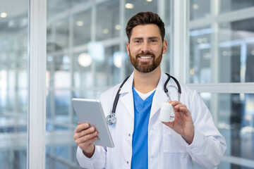 A smiling male doctor in a white coat holds a tablet and a medication bottle in a modern medical...