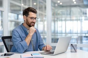 A professional man is working on a laptop, smiling, in a bright, modern office setting.