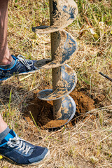 A person operates a handheld auger to drill a hole in the ground on a grassy field, preparing the...
