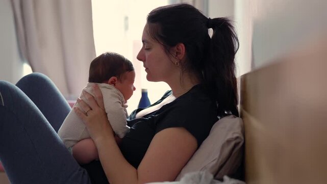 Mother kissing her baby before laying him down in bed and adjusting his posture. Warm daylight illuminates the scene, highlighting their intimate bond. Pink baby bassinet in the background