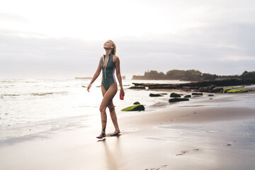 Woman walks gracefully on sand, earbuds in and camera in hand, smiling with eyes closed,...
