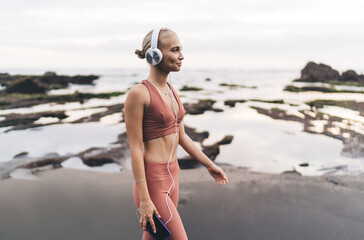 Young woman walks barefoot along the shore in activewear with headphones and phone, symbolizing...