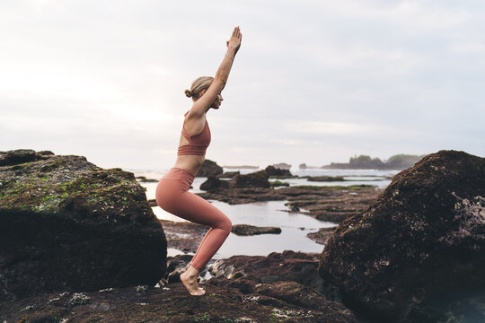 Side view of woman in activewear holding balance in chair pose between ocean rocks, showing inner concentration, breath control and focused elegance under cloudy morning sky.