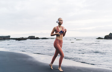 Woman jogs barefoot along shoreline, holding phone and listening with wired headphones, reflecting...