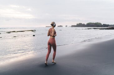 Back view of woman jogging toward horizon, black beach underfoot, headphones on, holding...