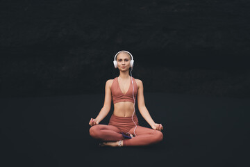 Young woman sits cross-legged on black sand in terracotta activewear with headphones, practicing...