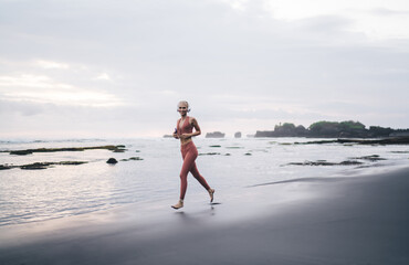 Smiling woman runs on volcanic beach holding phone and wearing headphones, expressing playful...