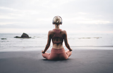 Back view of young woman meditating at shoreline in headphones, facing the ocean, practicing...