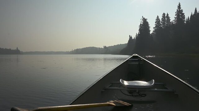 View from a canoe drifting on a calm lake covered in wildfire smoke with evergreen trees along the shore
