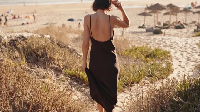 A young woman in a brown dress is walking on the sand towards the beach on a sunny day. She is walking away from the camera towards the water.