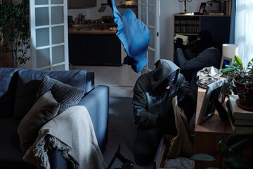 Group of masked men breaking into modern living room, searching through drawers and shelves,...