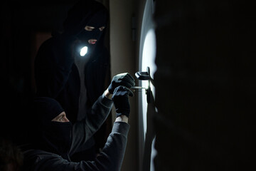 Two men wearing black balaclavas breaking into door with lock pick and flashlight, one crouching...